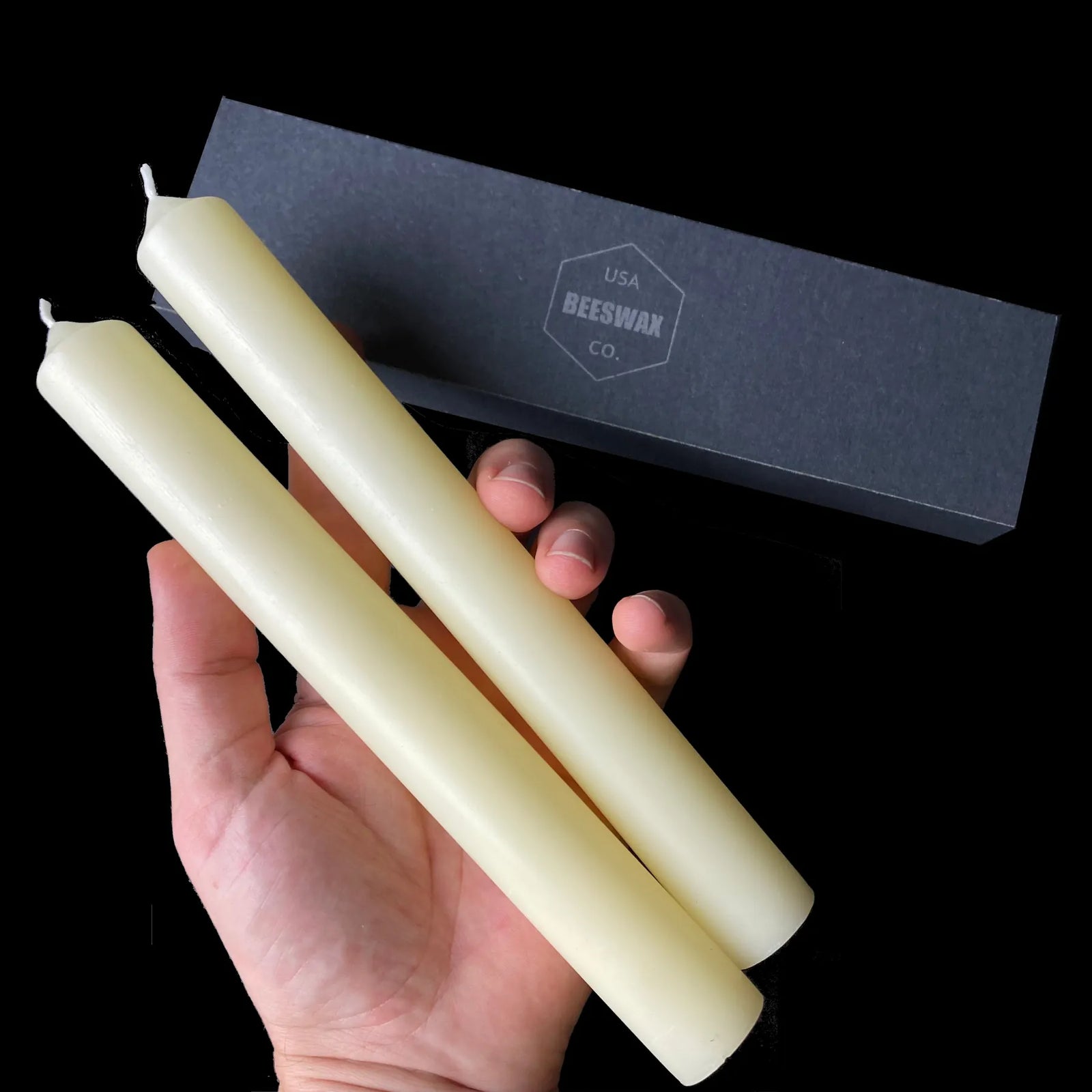 White beeswax taper candles, 1 inch wide, held in hand beside a branded gift box