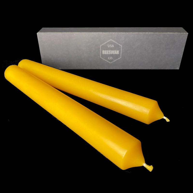 Two yellow dripless beeswax tapers laid on black with a gift box behind, 1 inch diameter