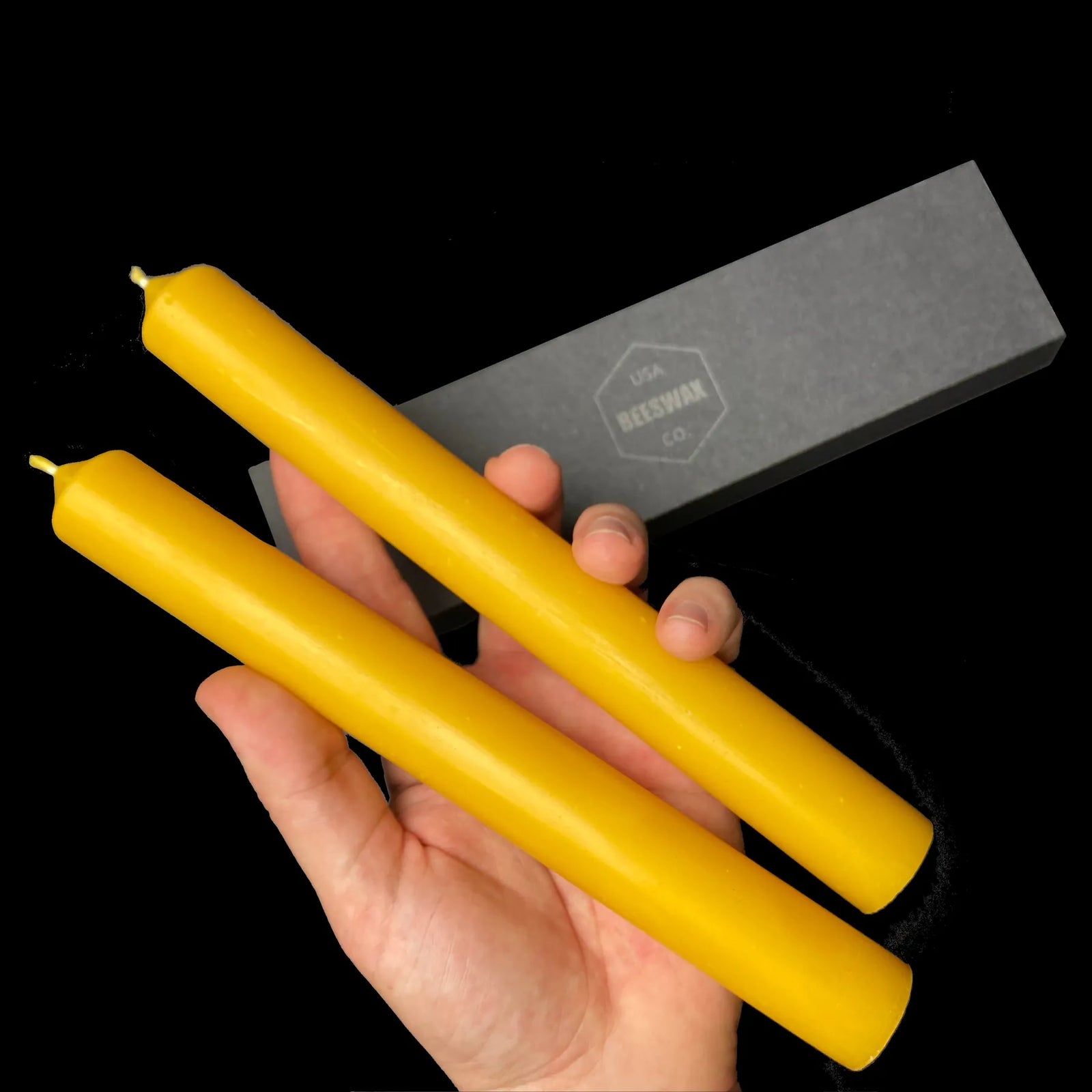 Yellow beeswax taper candles, 1 inch wide, held in hand beside a branded gift box